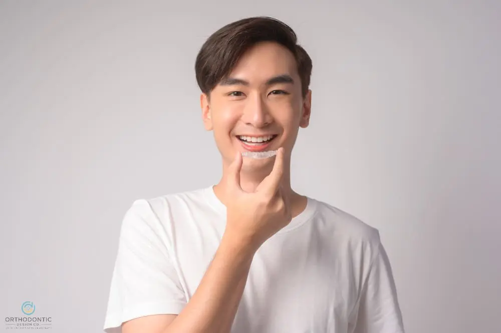 Smiling young man in a white t-shirt holds a clear aligner, promoting Orthodontic Design Co. Orthodontics in Madison and Huntsville, AL - How Does Invisalign Work in Madison and Huntsville, AL